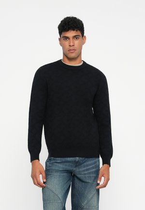Uomo che indossa un maglione nero a maniche lunghe con motivo e jeans in denim blu, in piedi contro uno sfondo bianco.