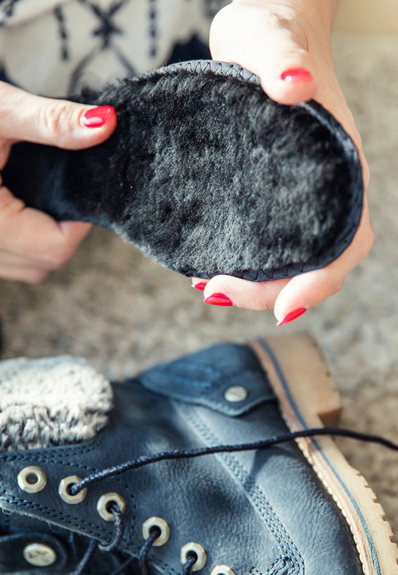 Black fur-lined insole held above a blue leather boot. Boot features a tan rubber sole, round eyelets, and gray fur trim at the ankle.