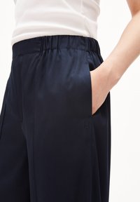 Osoba v námořnických modrých elastických kalhotách s rukou v boční kapse, doplněná bílým žebrovaným topem bez rukávů.