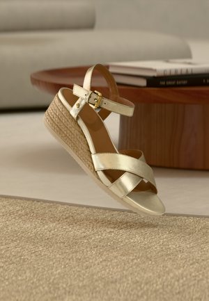 Sandalo zeppa beige con suola intrecciata in corda, cinturino alla caviglia con fibbia dorata e cinturini incrociati sulle dita dei piedi, mostrato vicino a un tavolo di legno con libri.