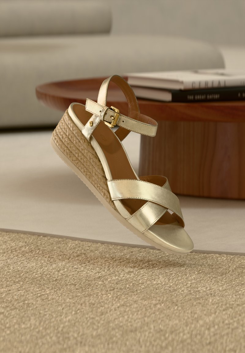 Μπεζ πλατφόρμα πέδιλο με πλεκτή σόλα από σχοινί, χρυσό πόρπη στον αστράγαλο και σταυρωτά λουράκια πάνω από τα δάχτυλα, τοποθετημένο κοντά σε ξύλινο τραπέζι με βιβλία.