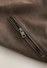 Fermeture éclair en métal partiellement ouverte sur un tissu marron lisse, montrant les détails de la couture le long des bords de la fermeture éclair.