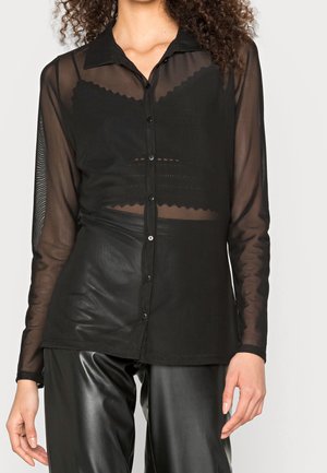 Femme portant un chemisier transparent noir à boutons sur un bralette à bords festonnés, assorti à un pantalon en cuir noir brillant, sur un fond uni.