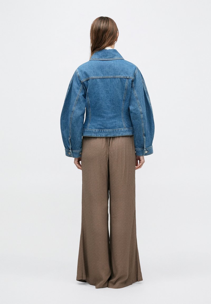 Denim jacket in blue with a fitted design, featuring puffed sleeves, a spread collar, and visible stitching. Paired with wide-legged brown pants.