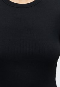Primo piano di una persona che indossa un top nero in tessuto a coste con scollo rotondo su uno sfondo chiaro uniforme.