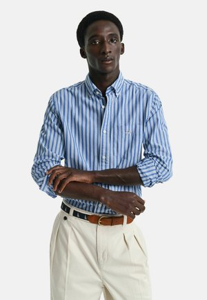 Blue and white striped button-down shirt with a chest pocket, paired with light beige pleated trousers and a brown belt with a nautical pattern.