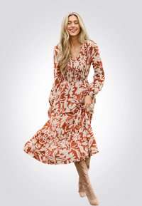 Maxikleid mit floralem Muster in Rost und Creme, mit gerüschtem Oberteil, langen Ärmeln und einem gestuften Rock. Kombiniert mit kniefreien Stiefeln.