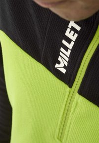 Fleece pullover with green and black color blocks, featuring a half-zip design and "Millet" logo in white at the neck. Textured fabric.