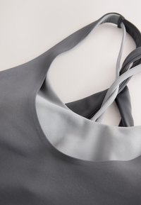 Grå atletisk tanktop med snoede skulderstropper lagt fladt på en hvid overflade, der viser detaljer ved halsudskæringen og stropperne.