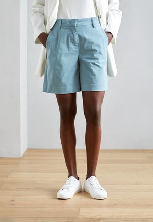 Persona con pantalones cortos azul claro, zapatillas blancas y una chaqueta blanca con las manos en los bolsillos, de pie sobre un suelo de madera.