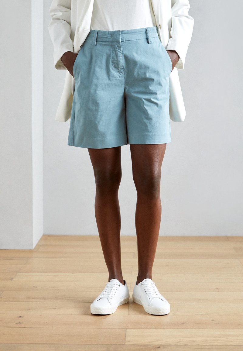Personne portant un short bleu clair, des baskets blanches et un blazer blanc, les mains dans les poches, debout sur un sol en bois.
