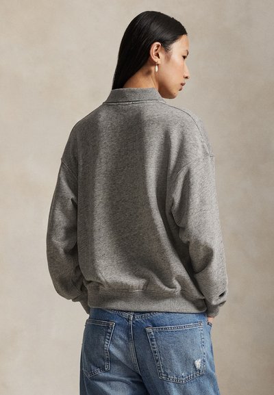 Pitkätukka nainen, jolla on helmikorvakorut, pukeutuneena harmaaseen puseroon ja sinisiin farkkuihin, seisoo sivuttain beigeä taustaa vasten.