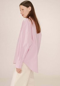 Camicia oversize a righe rosa chiaro con maniche lunghe e orlo asimmetrico, abbinata a pantaloni color crema. Il tessuto ha una texture sottile.