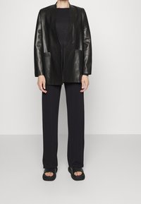 Černý kožený blejzr s jedním knoflíkem, dvěma předními kapsami a uvolněným střihem, kombinovaný s černými kalhotami s širokými nohavicemi a plochými sandály.