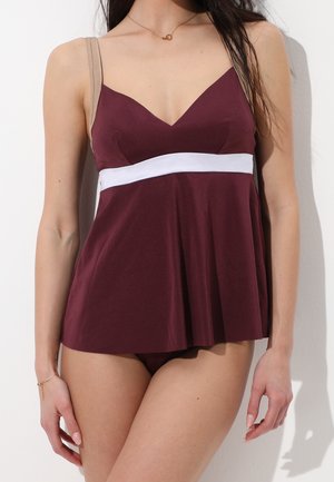 Donna che indossa un costume da bagno senza maniche color mogano con una fascia bianca sotto il seno e spalline beige, in piedi su uno sfondo bianco.