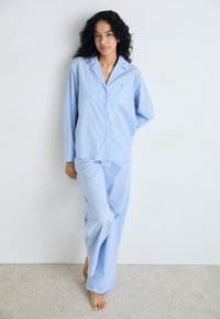 Ljusa blå rande pyjamas i mjukt tyg; knäppskjorta med krage och matchande löst sittande byxor. Inga anmärkningsvärda detaljer.