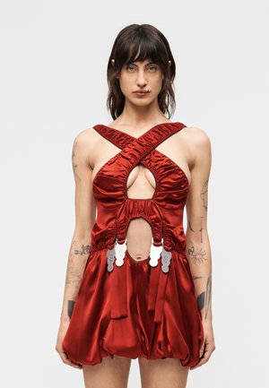 Femme aux cheveux foncés portant une robe rouge en satin sans manches avec des bretelles croisées, des découpes, des fermoirs décoratifs argentés et une jupe bouffante froncée, tatouages visibles.