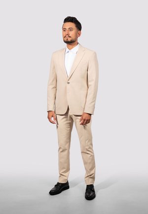 Beige Anzug aus strukturiertem Gewebe, mit einem Knopf, Reverskragen, zwei vorderen Taschen, kombiniert mit einem weißen Hemd und schwarzen Schuhen.