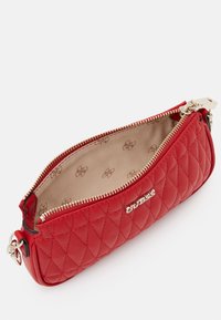 Bolso de mano acolchado rojo con cremallera y herrajes dorados. El interior presenta un forro beige liso con un sutil patrón de logotipos. Diseño compacto y redondeado.