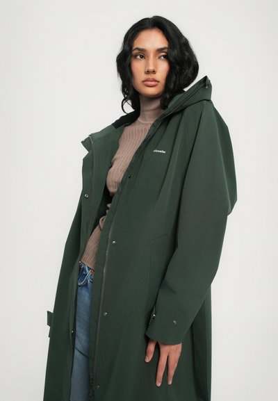 Veste imperméable verte avec fermeture éclair, capuche et poignets à bouton-pression, portée par-dessus un pull en maille beige et un jean bleu.