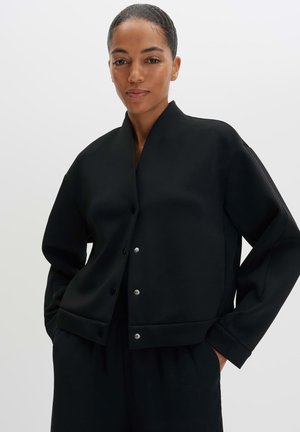 Frau mit Händen in den Taschen, die eine schwarze, geknöpfte Jacke mit hohem Kragen und langen Ärmeln vor einem einfarbigen hellen Hintergrund trägt.