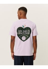 Lilac short-sleeve t-shirt featuring a green heart-shaped graphic with "LES DEUX LEGACY COURT" and an apple design on the back.