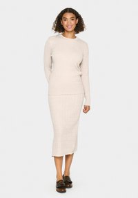 Dopasowany zestaw dzianinowy w jasnym beżu, składający się z dopasowanej bluzki z długim rękawem oraz spódnicy o długości midi z prążkowaną fakturą. Uzupełniony ciemnymi butami wsuwanymi.
