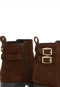 Brown suede ankle boots featuring a side zipper, two decorative belts with gold buckles, and a textured surface for added detail.