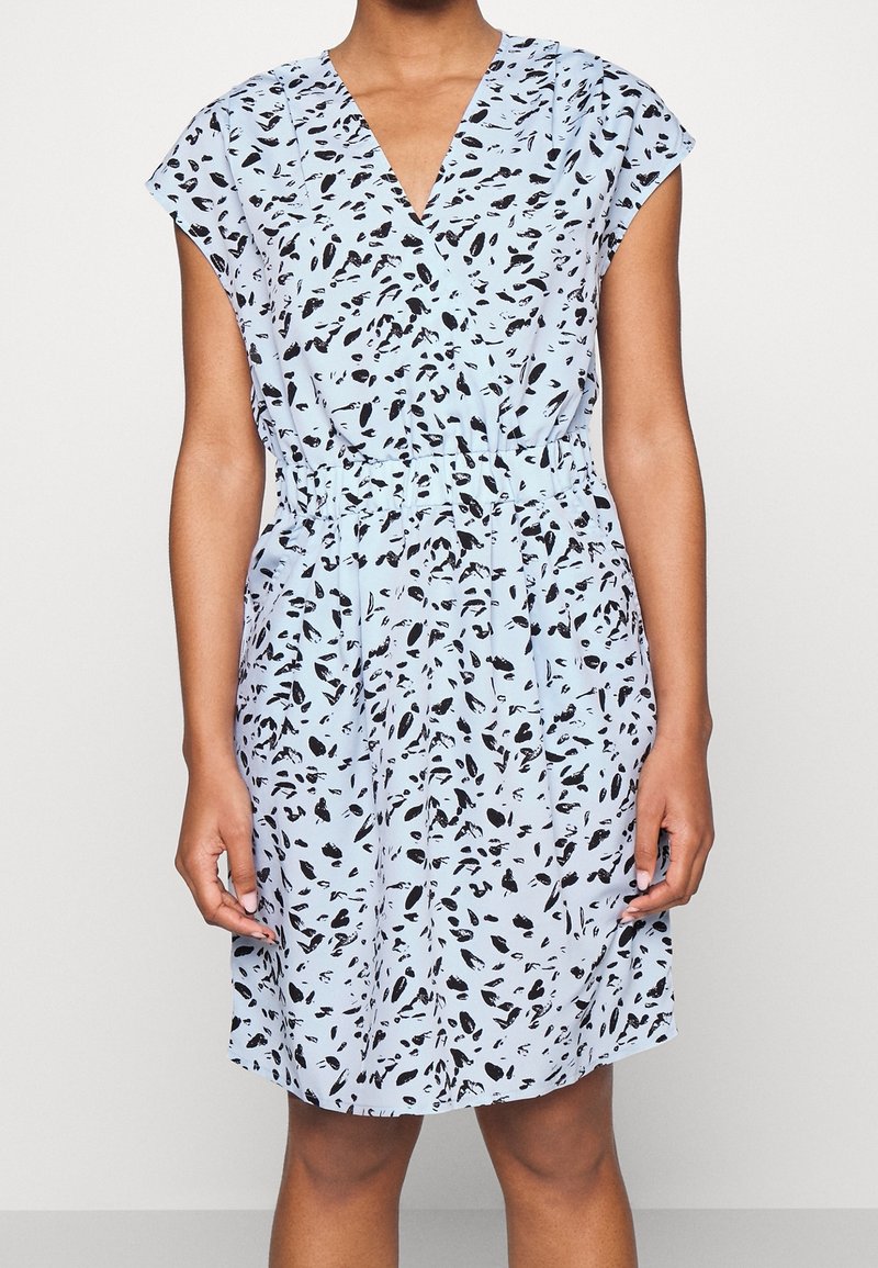 Femme portant une robe à manches courtes bleu clair avec des taches abstraites noires et une taille cintrée, se tenant devant un fond uni.