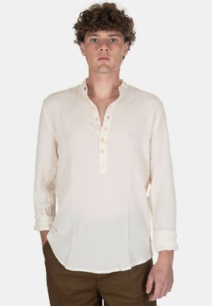 Uomo in piedi, rivolto in avanti, indossa una camicia color crema a maniche lunghe con colletto alla coreana e abbottonatura, e pantaloni marroni, mani lungo i fianchi.