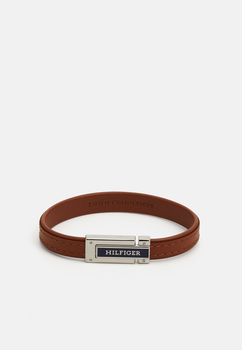 Pruun nahast käevõru, millel on hõbedane metallklamber, millel on ristkülikukujuline tumesinine aktsent, millel on kiri "HILFIGER". Tekstureeritud viimistlus ja õmbluste detailid.