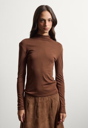 Woman with long dark hair wearing a brown long-sleeve top and a brown suede skirt, posing against a plain light background.