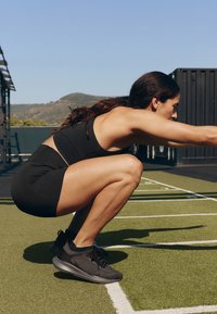 Femme en tenue de sport noire réalisant un exercice de squat profond sur une pelouse extérieure verte avec des montagnes et un ciel dégagé en arrière-plan.