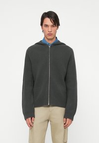 Felpa con zip grigio scuro a coste, dotata di cappuccio, con zip frontale, maniche lunghe e vestibilità comoda. Indossata sopra una camicia blu con colletto.