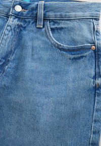 Gros plan sur la taille d'un jean en denim bleu, montrant le bouton, la passants de ceinture, la poche avant et les rivets en cuivre.