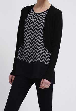 Cardigan nero su una maglietta grigia e blu navy a zigzag, con una vestibilità ampia e maniche lunghe, abbinato a pantaloni scuri.
