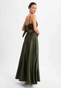 Olive green satin dress with a cutout back, adjustable straps, and a bow detail. Flowing silhouette with a smooth texture.