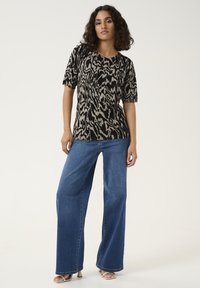 Top corto a maniche corte a motivi neri e beige con scollo rotondo, abbinato a jeans in denim blu a gamba larga e sandali aperti.