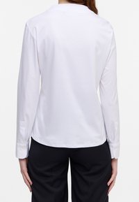 Camicia bianca a maniche lunghe con scollo rotondo, caratterizzata da una texture liscia e polsini con bottoni, abbinata a pantaloni scuri.