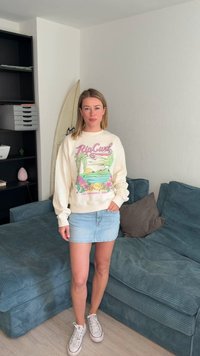 Mujer de pie en la sala de estar con un suéter ligero con estampado de playa, falda vaquera y zapatillas blancas, cerca de un sofá azul y una estantería negra.