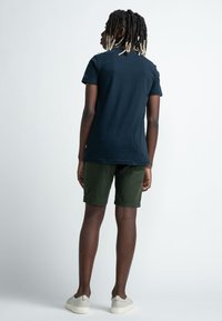 T-shirt di cotone blu navy con colletto arrotondato, abbinato a pantaloni corti verde oliva. La persona indossa scarpe slip-on grigio chiaro.