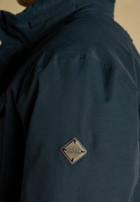 Detailný záber na rukáv tmavomodrej bundy so štvorcovou koženou nášivkou s embossovaným názvom značky "Joules" v zlatej farbe.