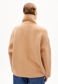 Beige fleece pullover met een hoge kraag, verlaagde schouders en een relaxte pasvorm, met een zachte textuur en stikdetails op de rug.