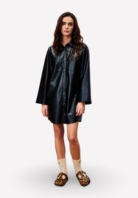 Robe-chemise en similicuir noir avec fermeture boutonnée sur le devant, manches longues et deux poches poitrine, associée à des chaussures imprimé léopard et des chaussettes crème.