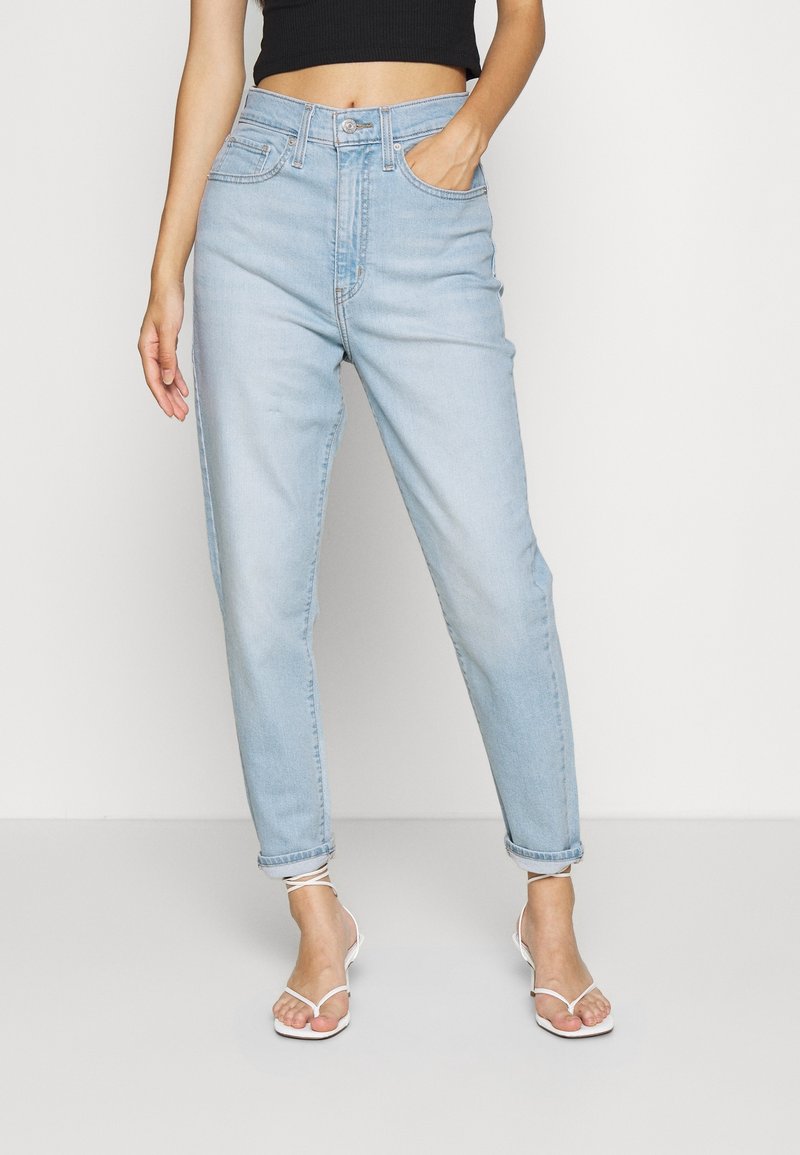 Jean taille haute bleu clair avec jambes fuselées, présentant un design à cinq poches, des passants de ceinture et des ourlets retroussés. Porté avec des sandales blanches à brides.