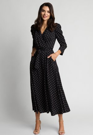 Kobieta o ciemnych włosach ubrana w czarną sukienkę midi w beżowe groszki, z bufkami na ramionach, paskiem w talii oraz beżowymi sandałami na wysokim obcasie, uśmiechająca się.