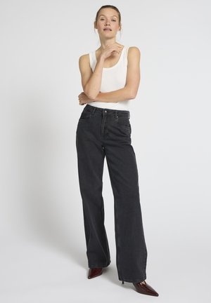 Sieviete stāv, šķērsām salikusi vienu roku un pieskārusi zodu, valkā baltu bezroku topu, melnas augsta vidukļa plata gurnu džinsas un bordo krāsas smailas kurpes ar augstu papēdi.