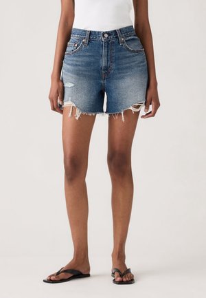 Kvinde iført denimshorts med frynset kant, hvid top og sorte flipflops, står mod en ensfarvet lys baggrund.
