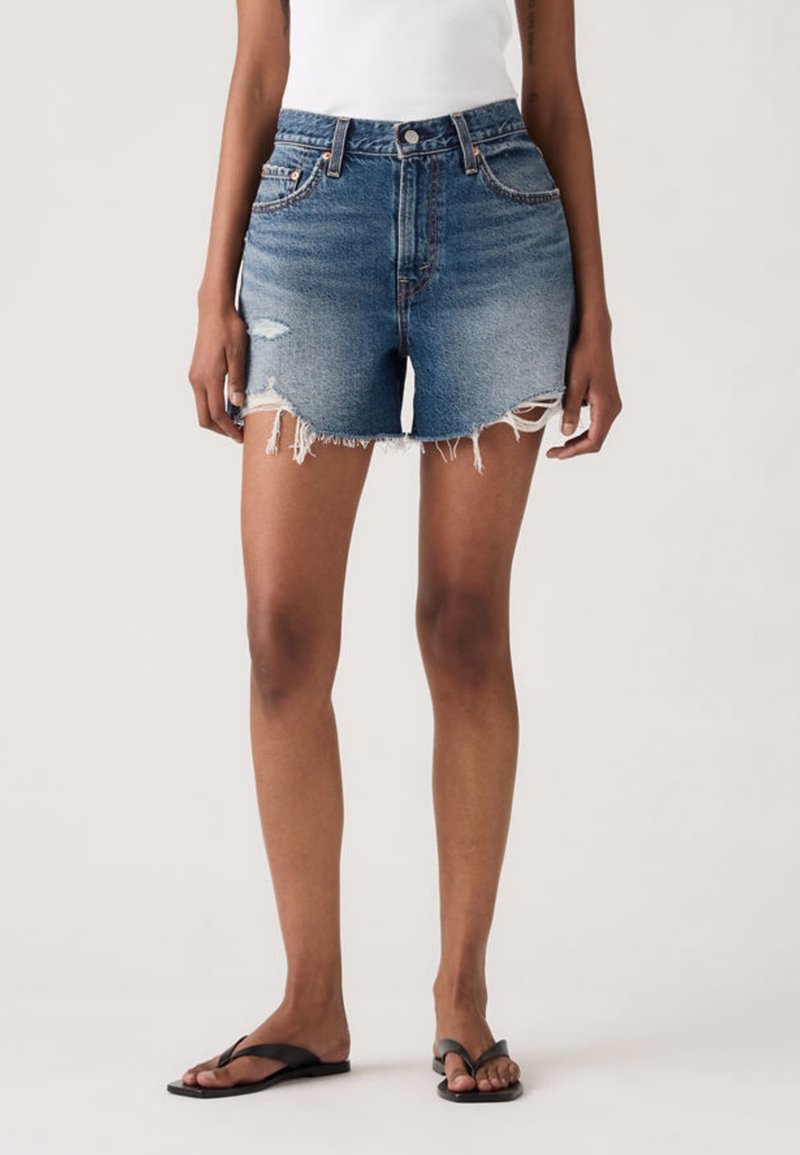 Femme portant un short en jean à ourlet effiloché, un haut blanc et des tongs noires, debout devant un fond clair uni.