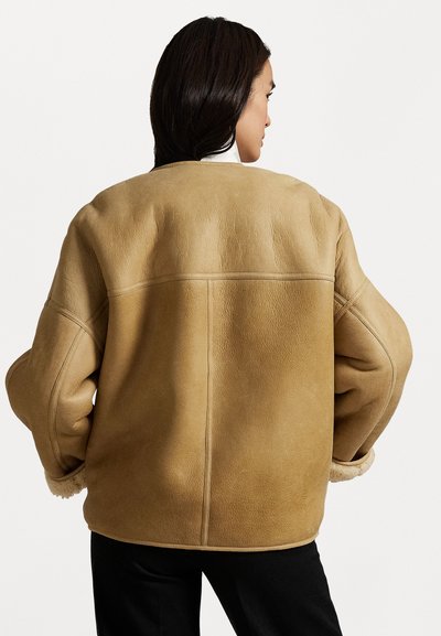 Giubbotto shearling color beige con vestibilità ampia, cuciture distintive e un esterno texturizzato. Le maniche a polsino rivelano un morbido rivestimento interno.